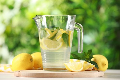 Photo of Refreshing lemonade with mint in jug among fruits on light table against blurred green background, closeup