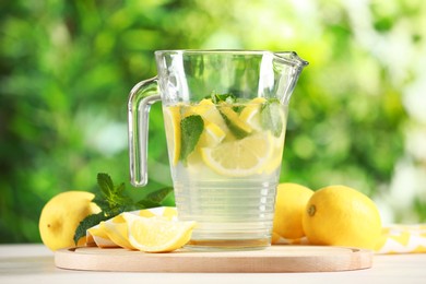 Refreshing lemonade with mint in jug among fruits on light table against blurred green background, closeup