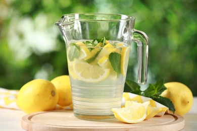 Refreshing lemonade with mint in jug among fruits on light table against blurred green background, closeup