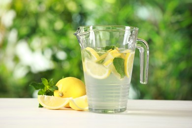 Refreshing lemonade with mint in jug on light table against blurred green background