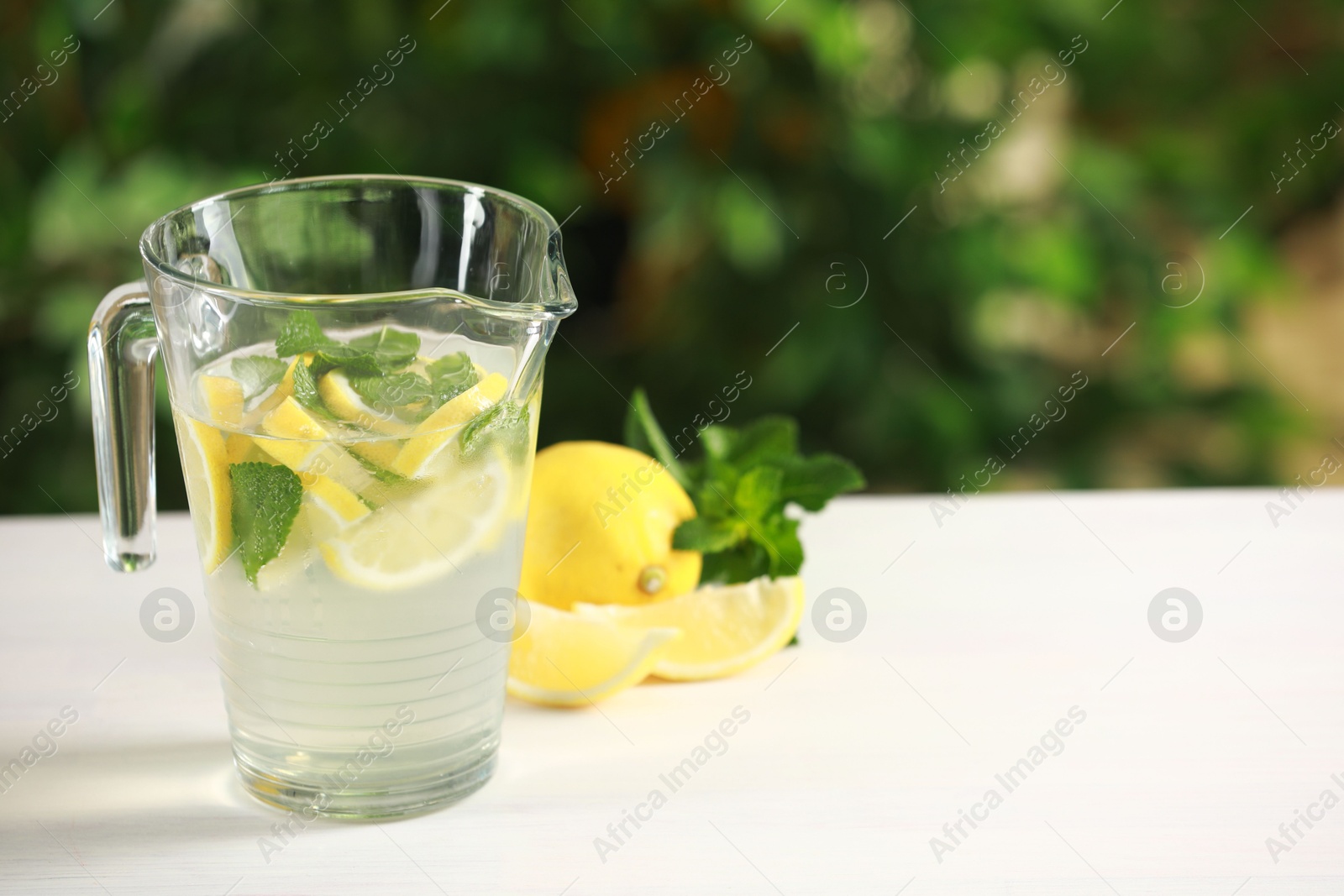 Photo of Refreshing lemonade with mint in jug on light table against blurred green background. Space for text