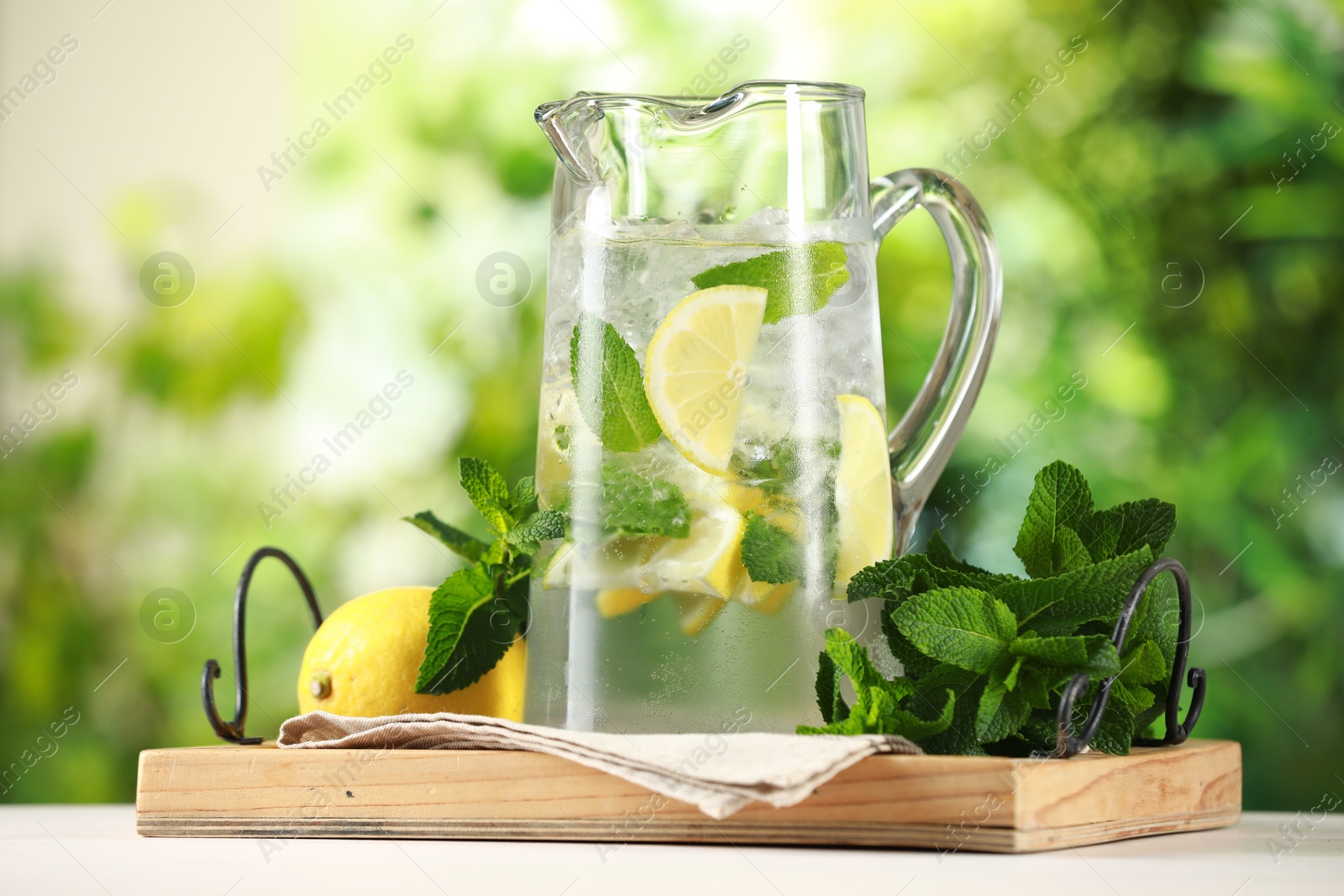 Photo of Refreshing lemonade with mint in jug and citrus fruit on light table against blurred green background