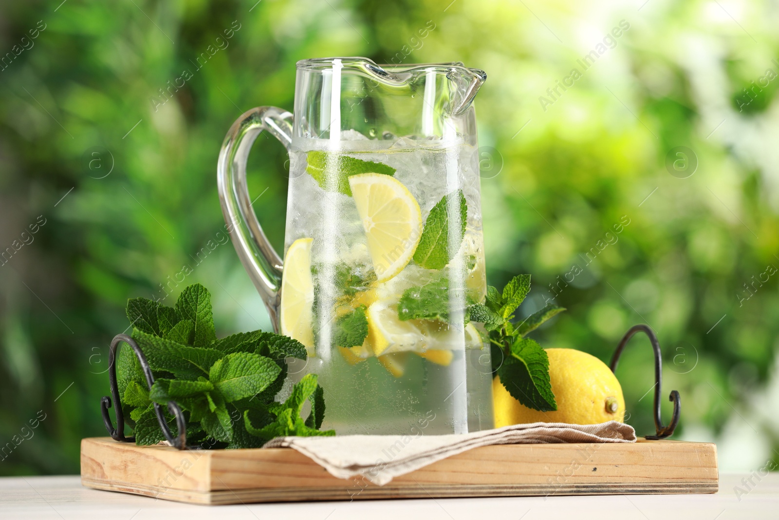 Photo of Refreshing lemonade with mint in jug and citrus fruit on light table against blurred green background