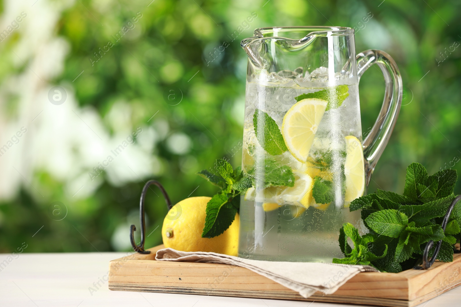 Photo of Refreshing lemonade with mint in jug and citrus fruit on light table against blurred green background. Space for text