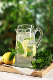 Photo of Refreshing lemonade with mint in jug and citrus fruit on light table against blurred green background