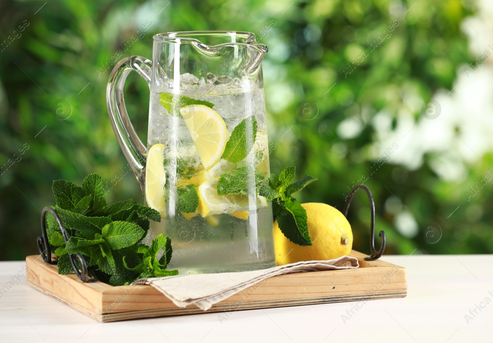 Photo of Refreshing lemonade with mint in jug and citrus fruit on light table against blurred green background, closeup