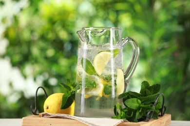 Photo of Refreshing lemonade with mint in jug and citrus fruit on table against blurred green background