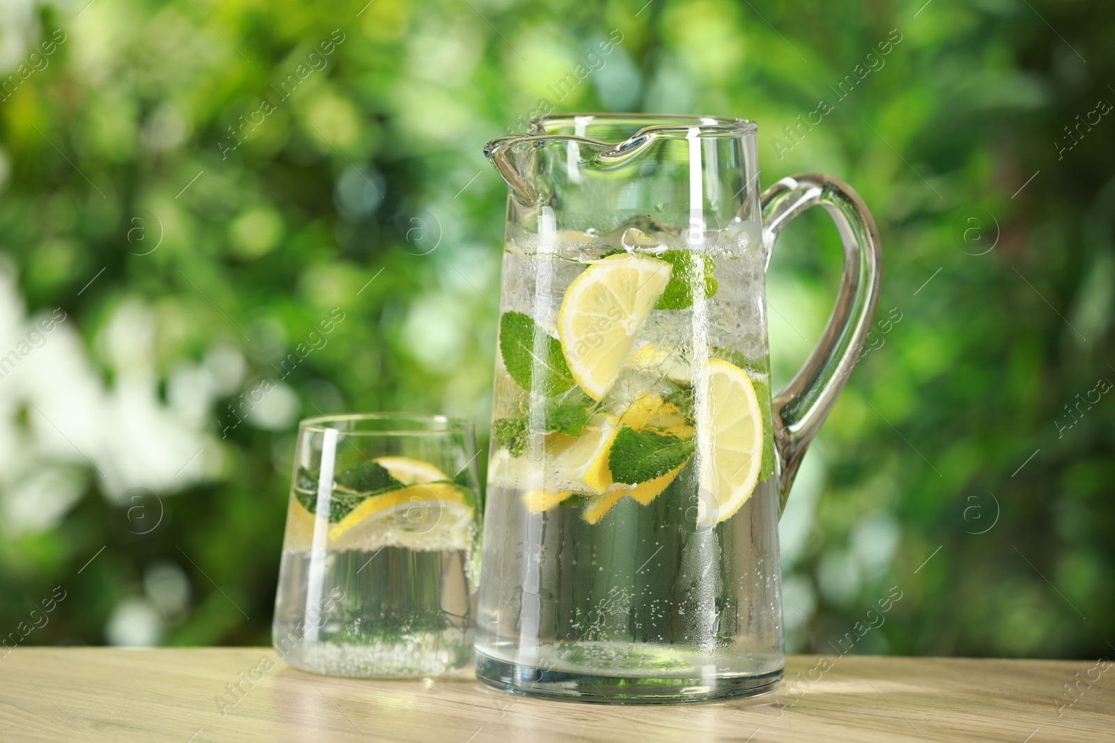 Photo of Refreshing lemonade with mint in jug and glass on wooden table against blurred green background