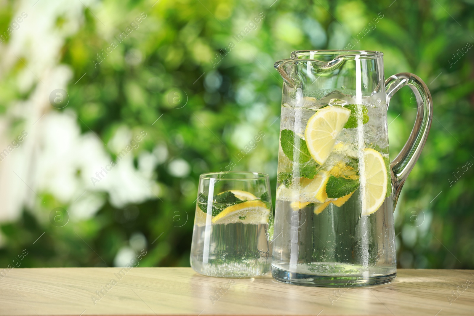 Photo of Refreshing lemonade with mint in jug and glass on wooden table against blurred green background. Space for text