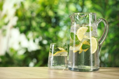 Refreshing lemonade with mint in jug and glass on wooden table against blurred green background. Space for text