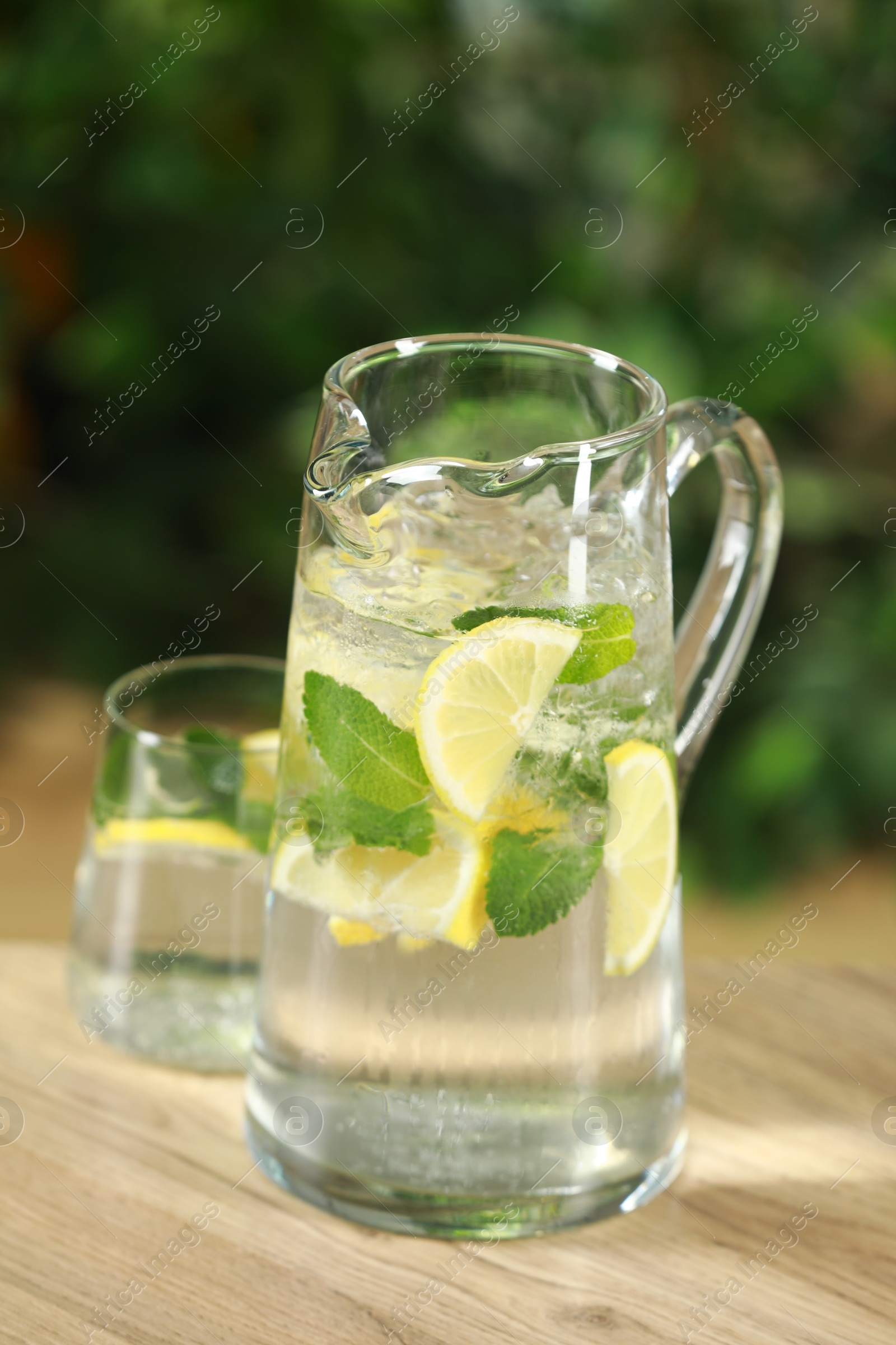 Photo of Refreshing lemonade with mint in jug and glass on wooden table against blurred green background, closeup