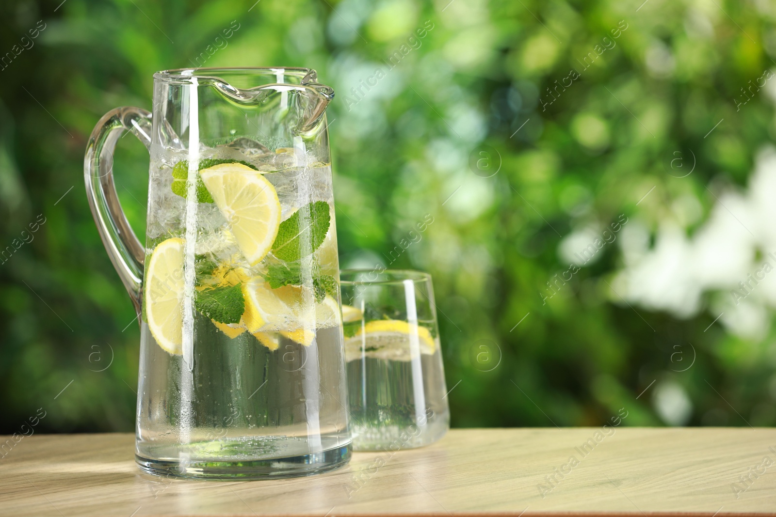 Photo of Refreshing lemonade with mint in jug and glass on wooden table against blurred green background. Space for text