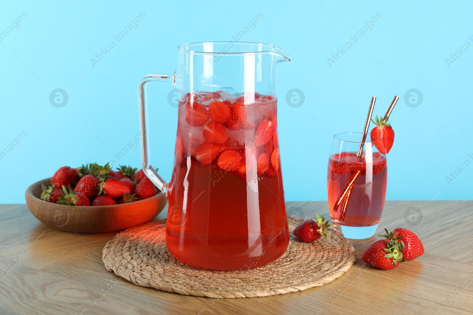 Photo of Tasty strawberry lemonade in jug, glass and berries on wooden table against light blue background