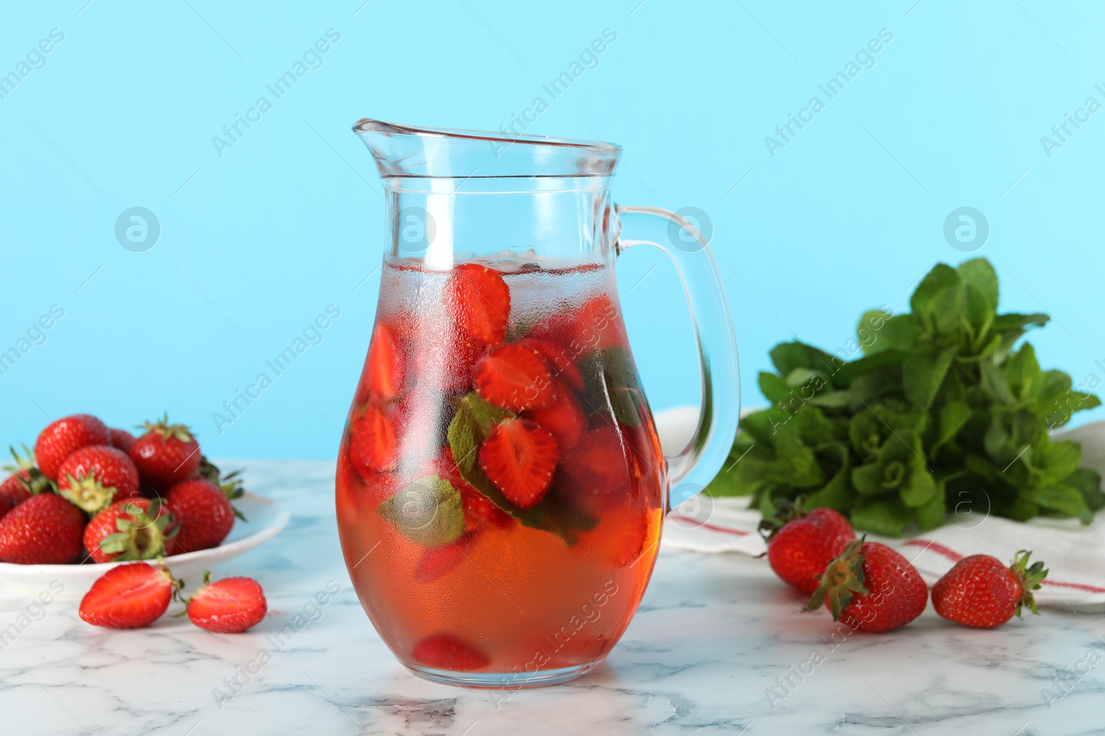 Photo of Tasty strawberry lemonade with mint in jug and berries on white marble table against light blue background