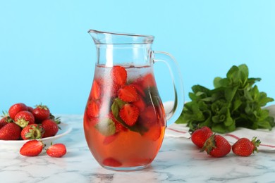 Tasty strawberry lemonade with mint in jug and berries on white marble table against light blue background