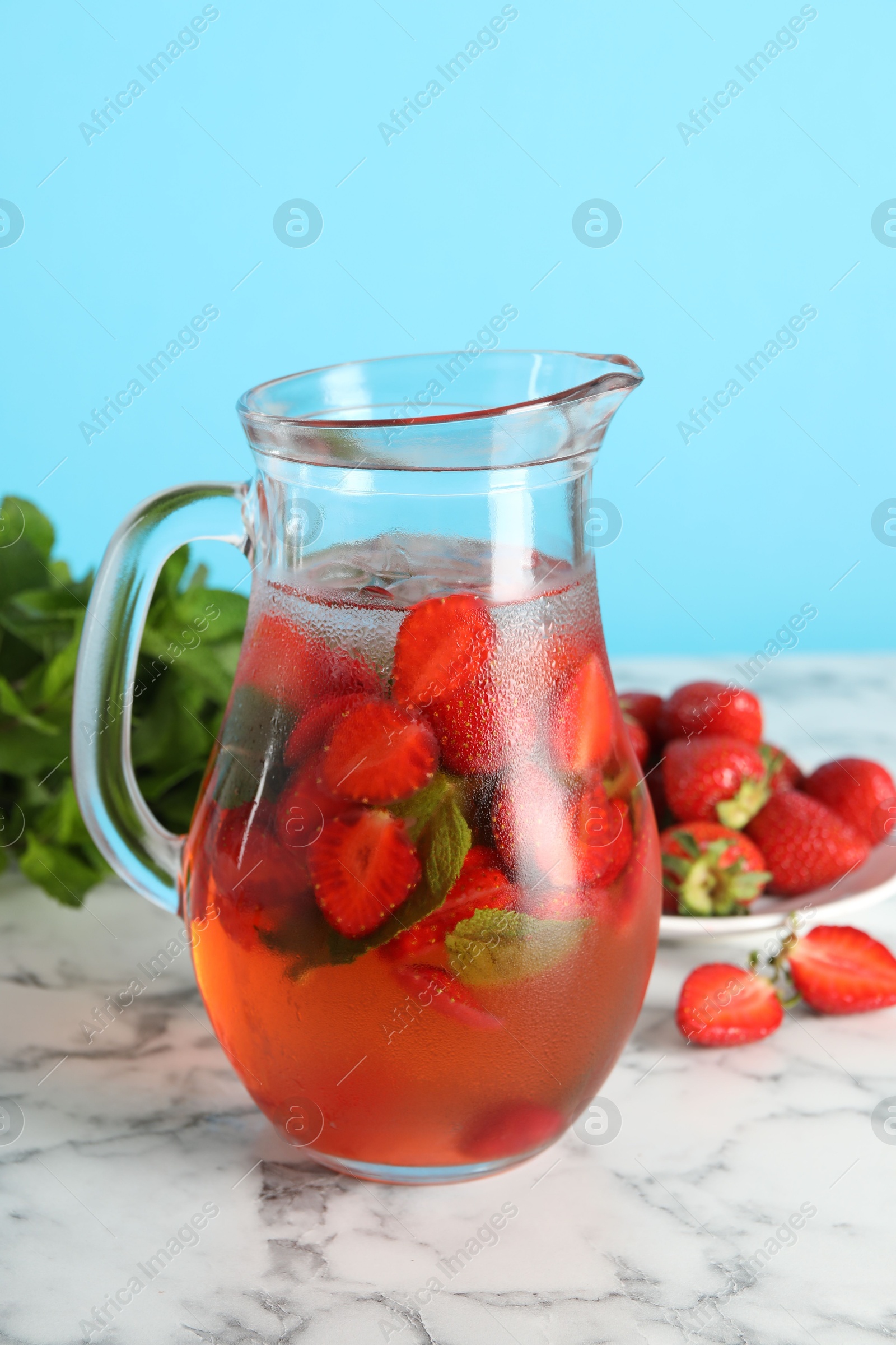 Photo of Tasty strawberry lemonade with mint in jug and berries on white marble table against light blue background