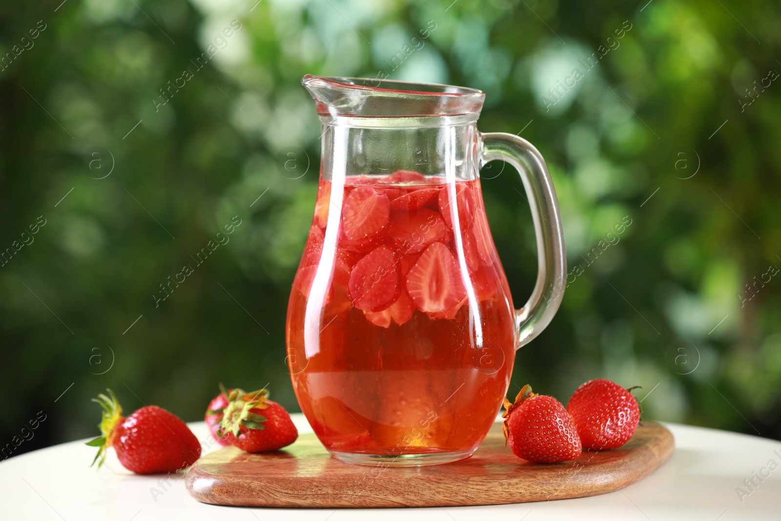 Photo of Tasty strawberry lemonade in jug and berries on white table against blurred green background