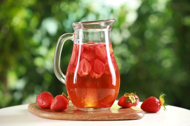 Photo of Tasty strawberry lemonade in jug and berries on white table against blurred green background