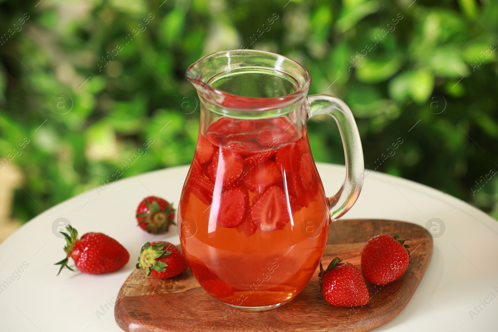 Photo of Tasty strawberry lemonade in jug and berries on white table against blurred green background, closeup