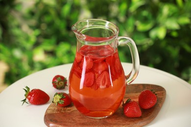 Tasty strawberry lemonade in jug and berries on white table against blurred green background, closeup