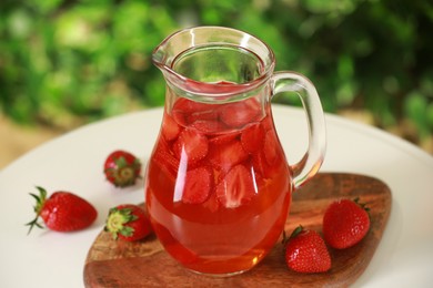 Photo of Tasty strawberry lemonade in jug and berries on white table against blurred green background, closeup