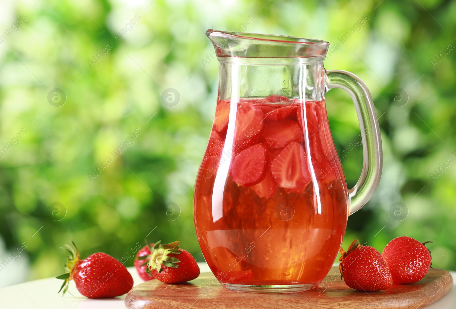 Photo of Tasty strawberry lemonade in jug and berries on white table against blurred green background, closeup. Space for text