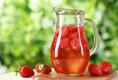 Tasty strawberry lemonade in jug and berries on white table against blurred green background, closeup. Space for text