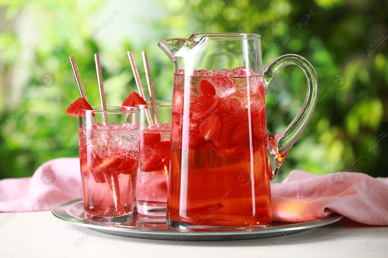 Photo of Tasty strawberry lemonade in jug and glasses on white table against blurred green background