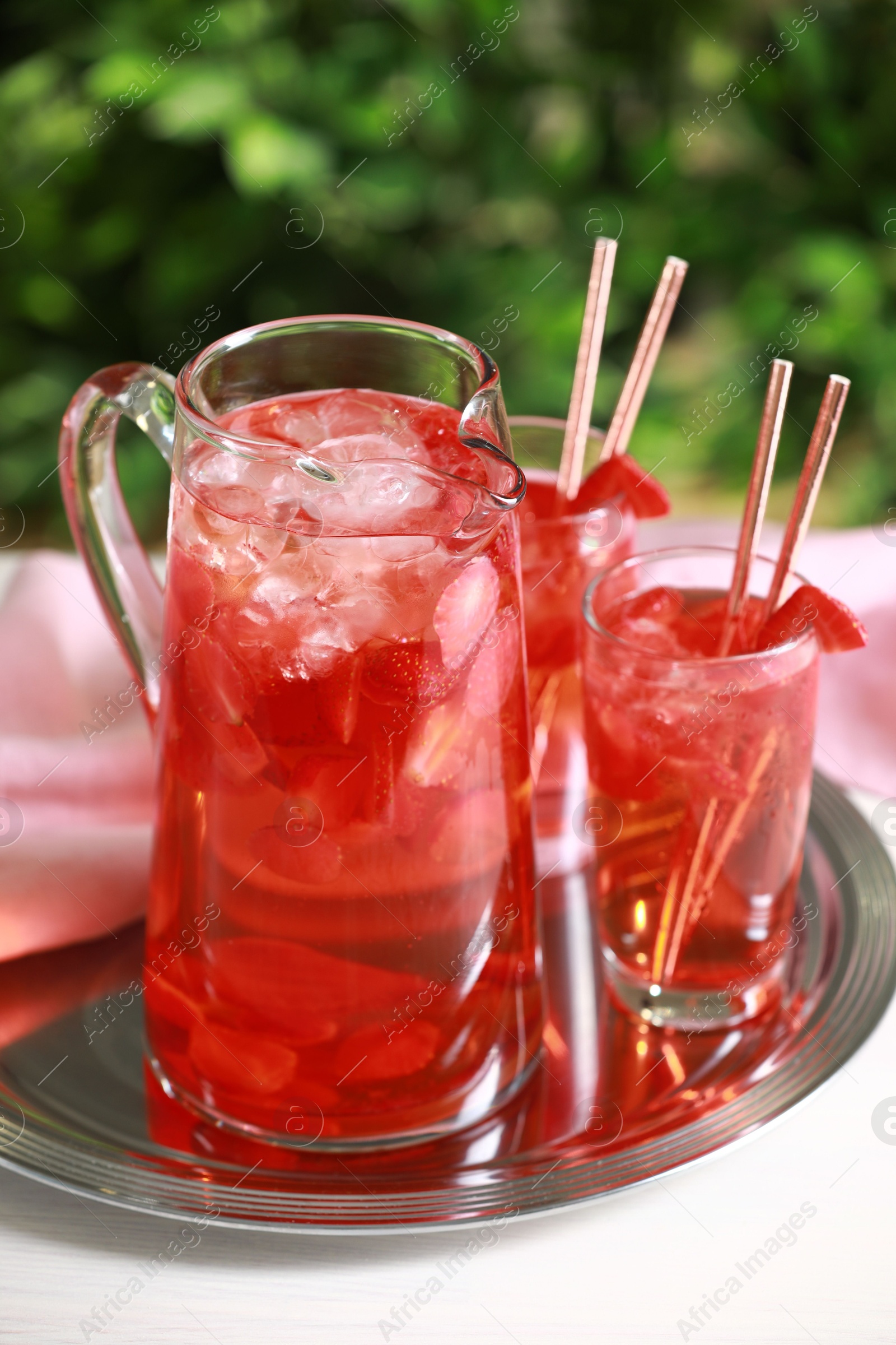 Photo of Tasty strawberry lemonade in jug and glasses on white table against blurred green background