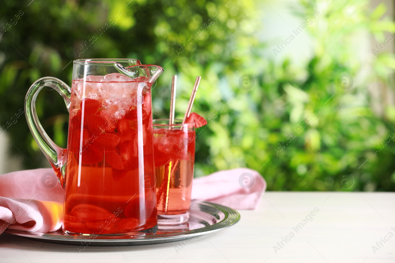 Photo of Tasty strawberry lemonade in jug and glasses on white table against blurred green background. Space for text