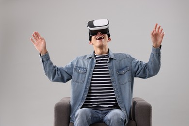 Emotional young man with virtual reality headset sitting on armchair against light grey background