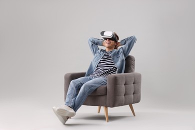 Happy young man with virtual reality headset sitting on armchair against light grey background