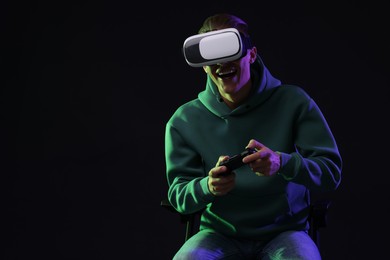 Happy young man with virtual reality headset and controller sitting on chair in neon lights against black background, space for text