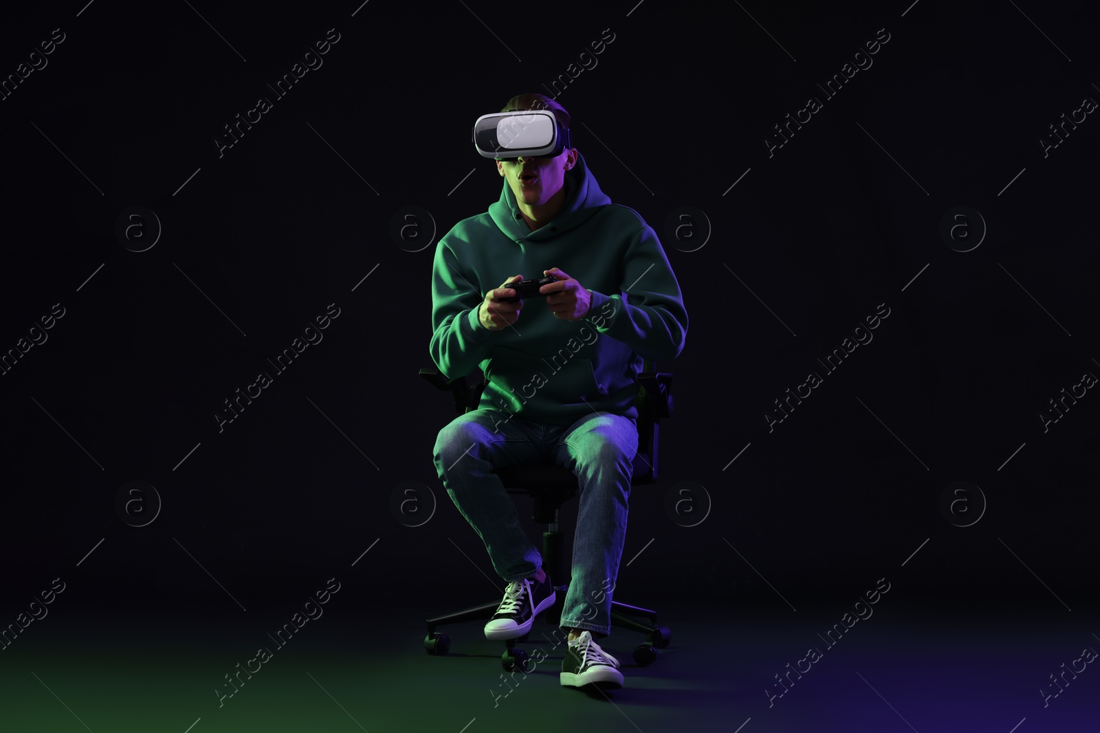 Photo of Emotional young man with virtual reality headset and controller sitting on chair in neon lights against black background