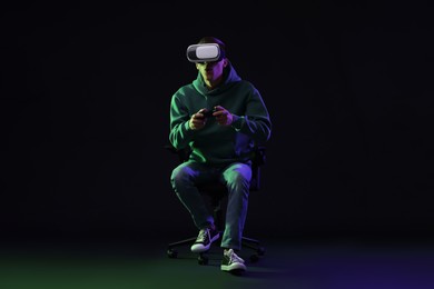 Photo of Emotional young man with virtual reality headset and controller sitting on chair in neon lights against black background