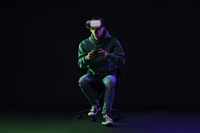 Emotional young man with virtual reality headset and controller sitting on chair in neon lights against black background