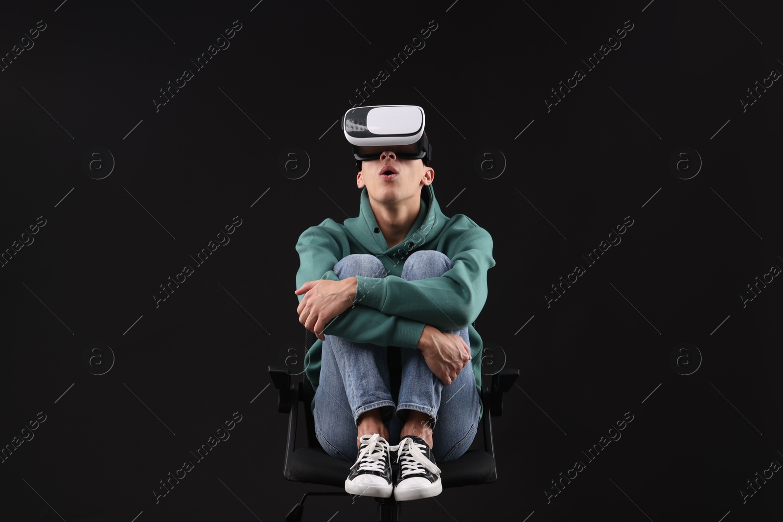 Photo of Emotional young man with virtual reality headset sitting on chair against black background