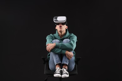 Emotional young man with virtual reality headset sitting on chair against black background