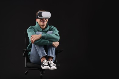 Photo of Happy young man with virtual reality headset sitting on chair against black background, space for text