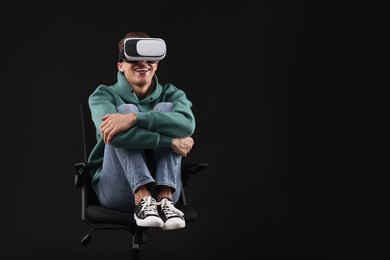 Happy young man with virtual reality headset sitting on chair against black background, space for text