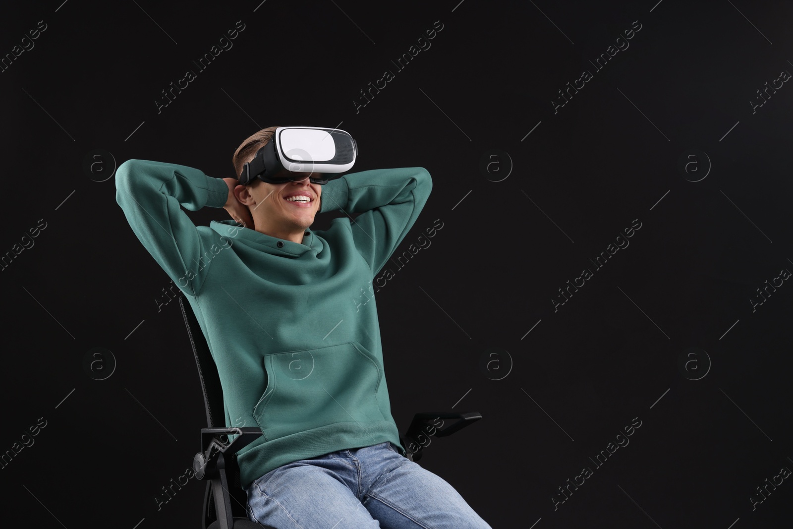 Photo of Happy young man with virtual reality headset sitting on chair against black background, space for text