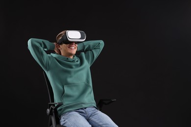 Happy young man with virtual reality headset sitting on chair against black background, space for text