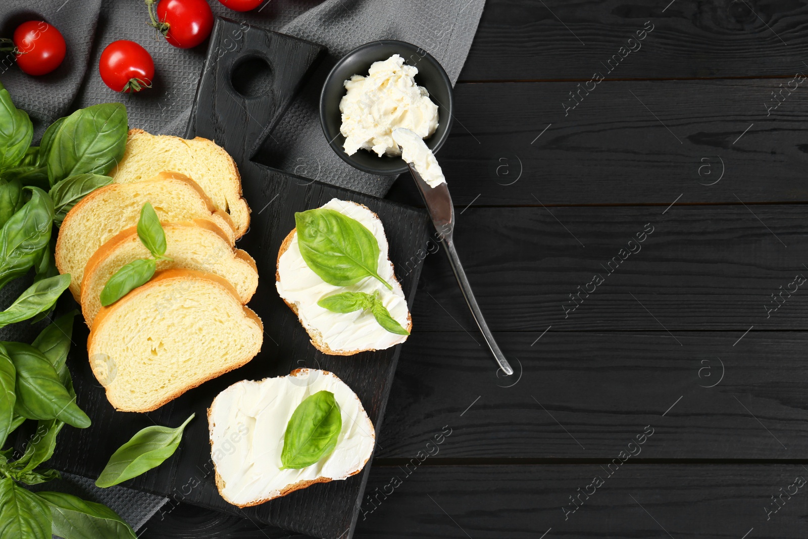 Photo of Pieces of bread with cream cheese and basil served on black wooden table, flat lay. Space for text