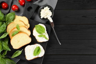 Pieces of bread with cream cheese and basil served on black wooden table, flat lay. Space for text