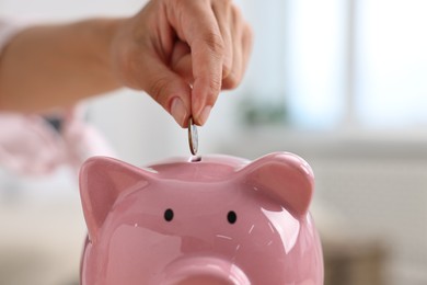 Photo of Woman putting coin into pink piggy bank at home, closeup