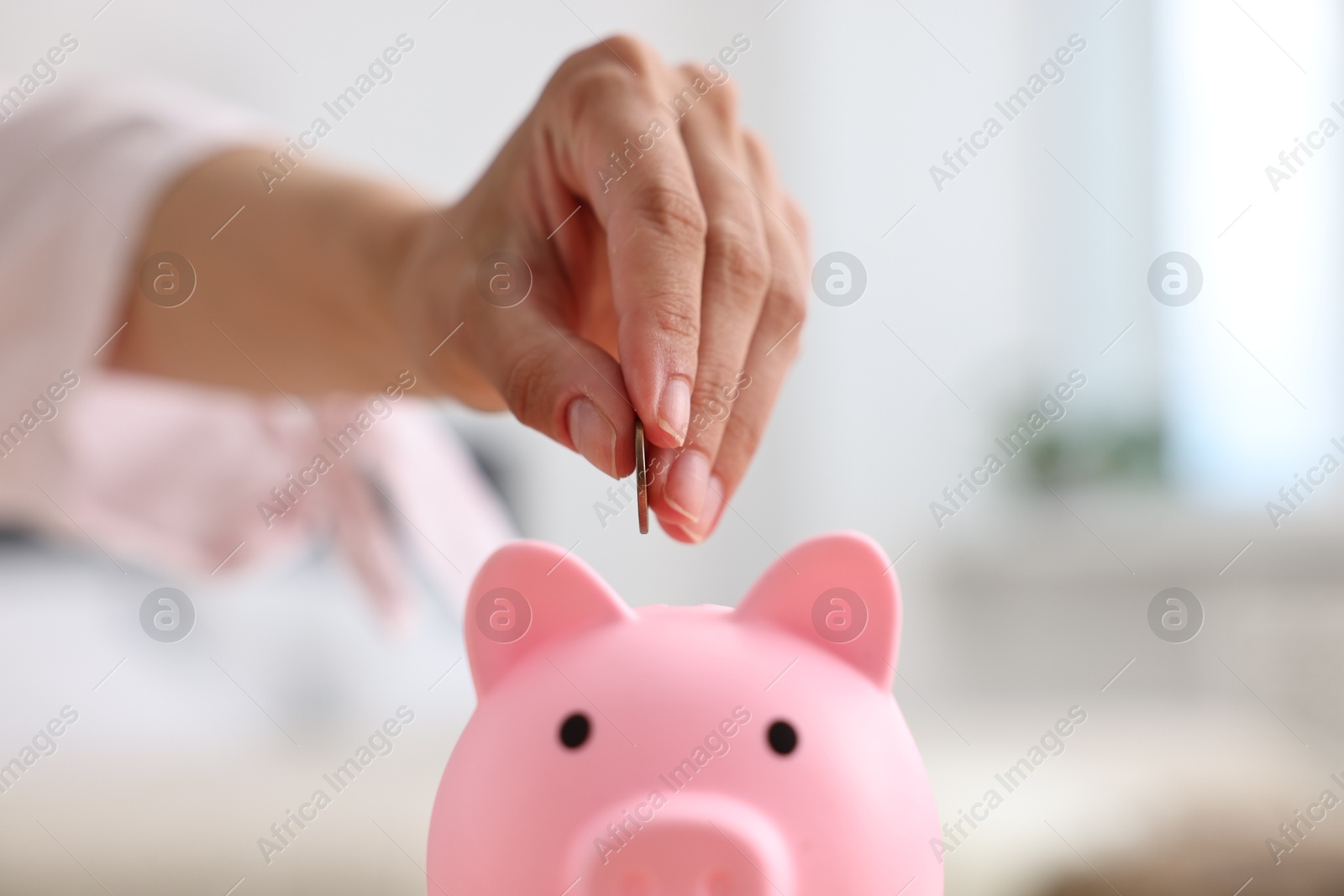 Photo of Woman putting coin into pink piggy bank at home, closeup