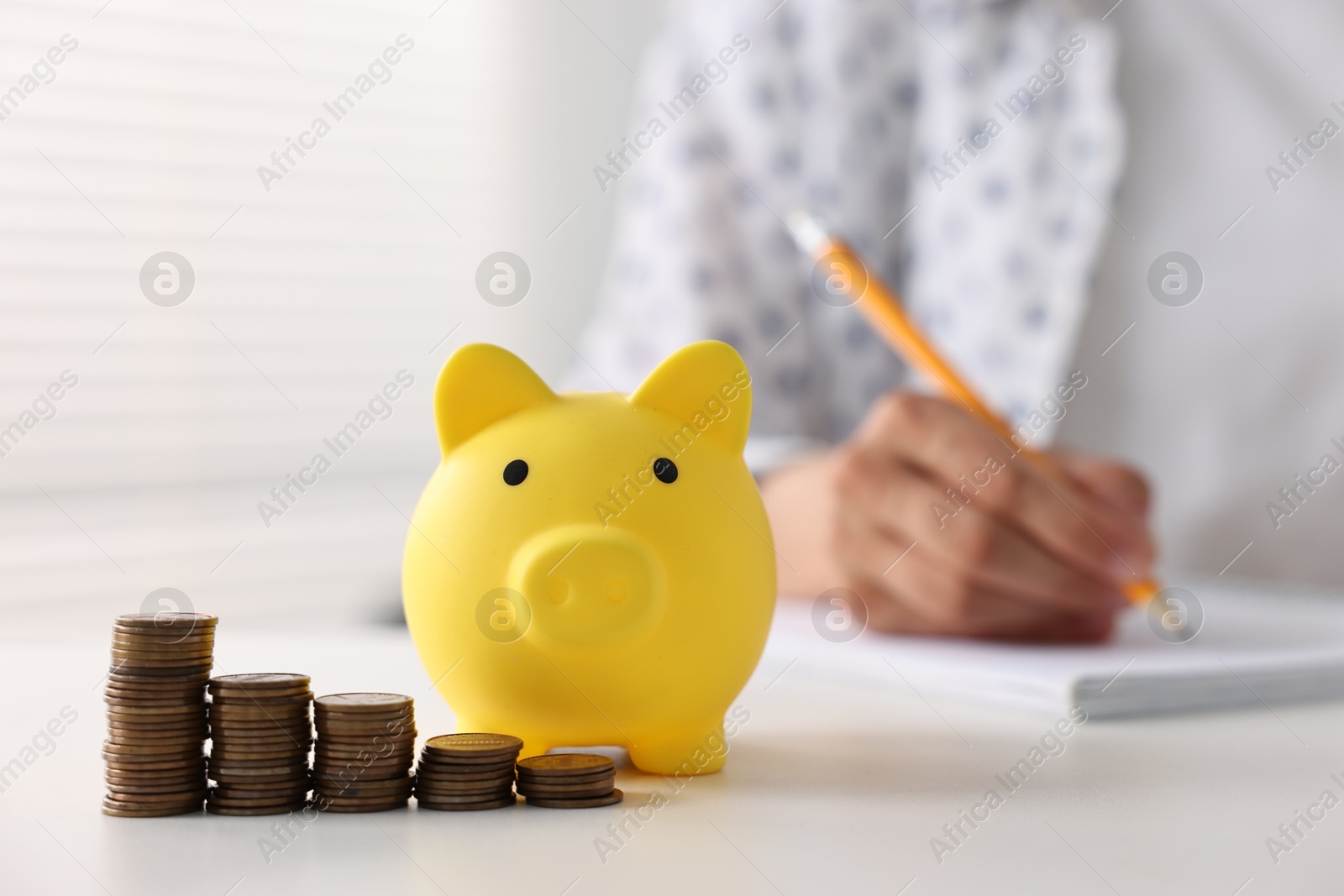 Photo of Woman taking notes at table indoors, focus on piggy bank and coins