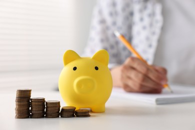 Photo of Woman taking notes at table indoors, focus on piggy bank and coins