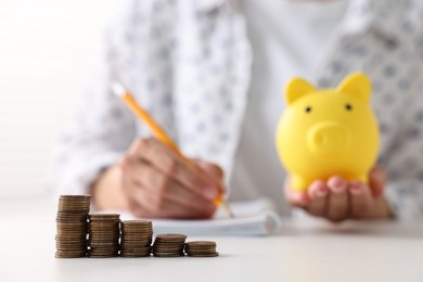 Photo of Woman with piggy bank taking notes at table indoors, focus on coins