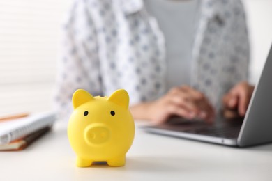 Woman using laptop at white table indoors, focus on piggy bank